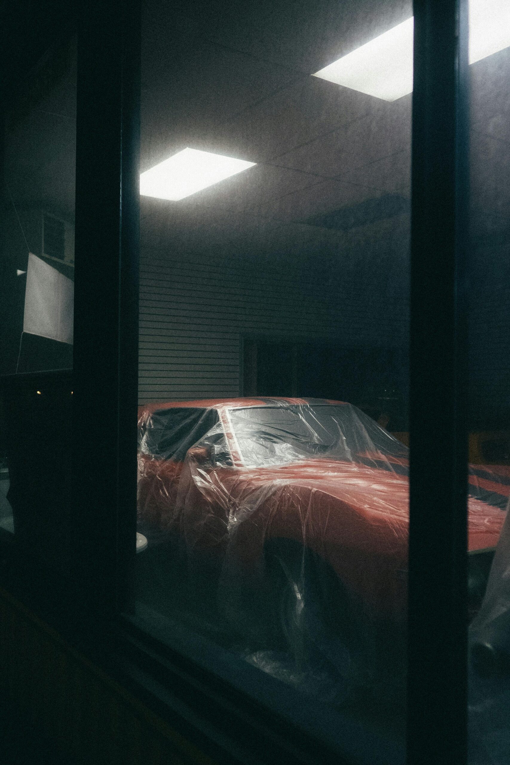 A dimly lit indoor scene showcasing a classic red car covered with protective plastic.
